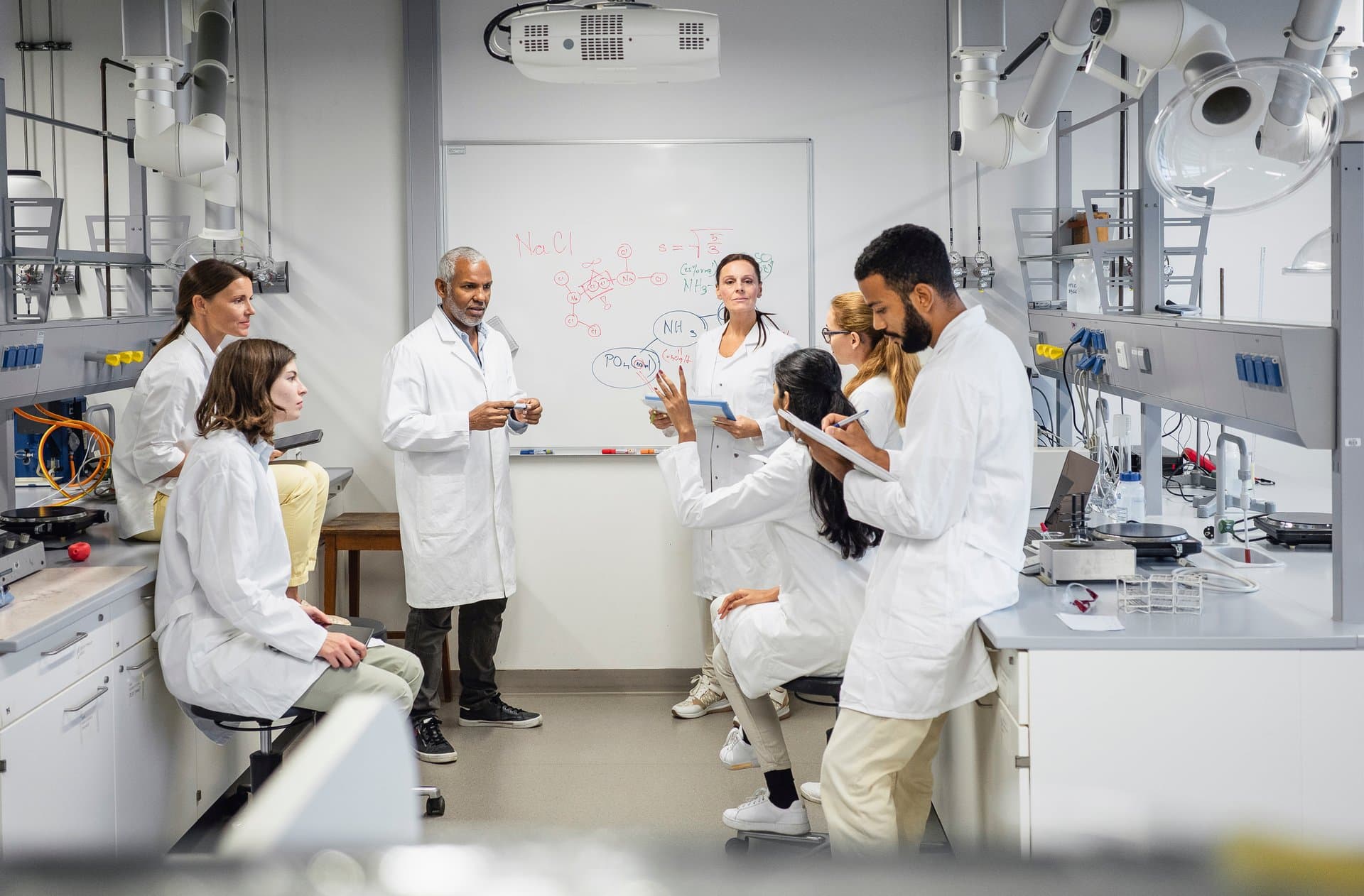 Group of scientists in white coats discussing chemical formulas on a whiteboard during a research meeting in a laboratory.