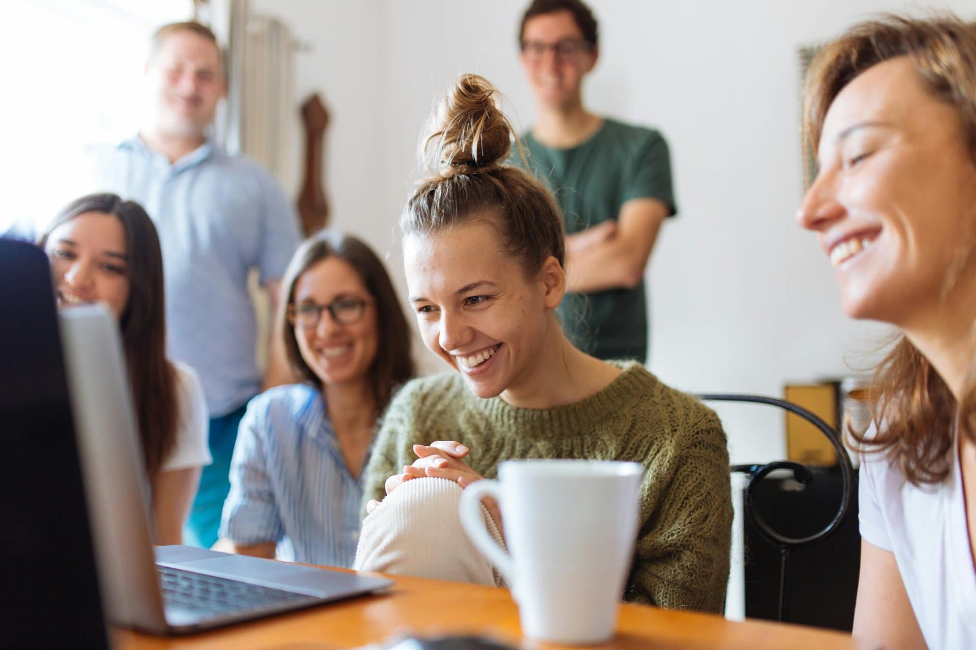 Diverse group of young adults laughing together while looking at a laptop screen, sharing coffee in a bright, casual indoor setting.