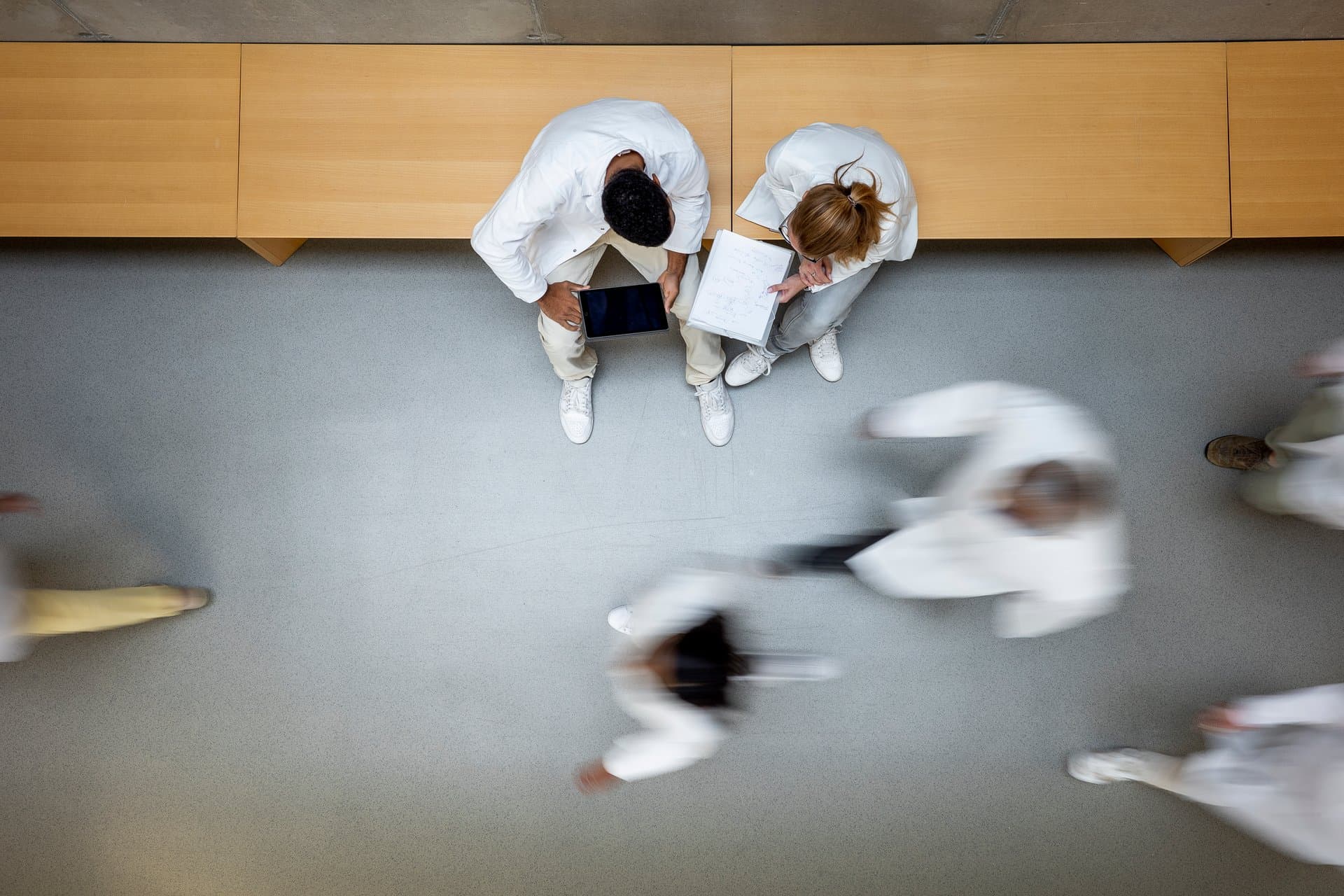 Aerial view of two clinicians in white coats reviewing data on a tablet and notes while colleagues walk briskly through a hospital hallway.