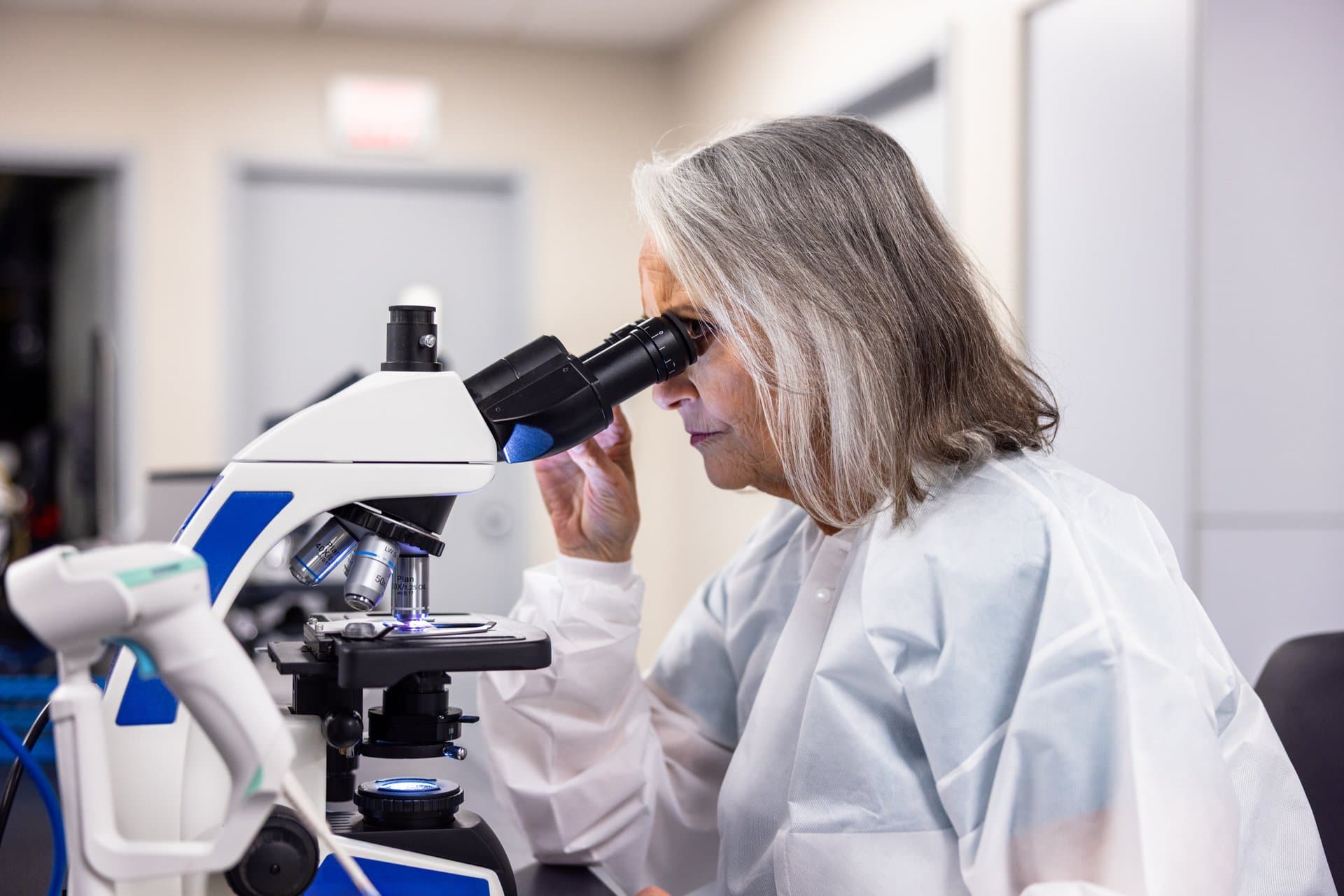 Older female researcher in white lab coat examining specimen through microscope in clinical research laboratory setting.