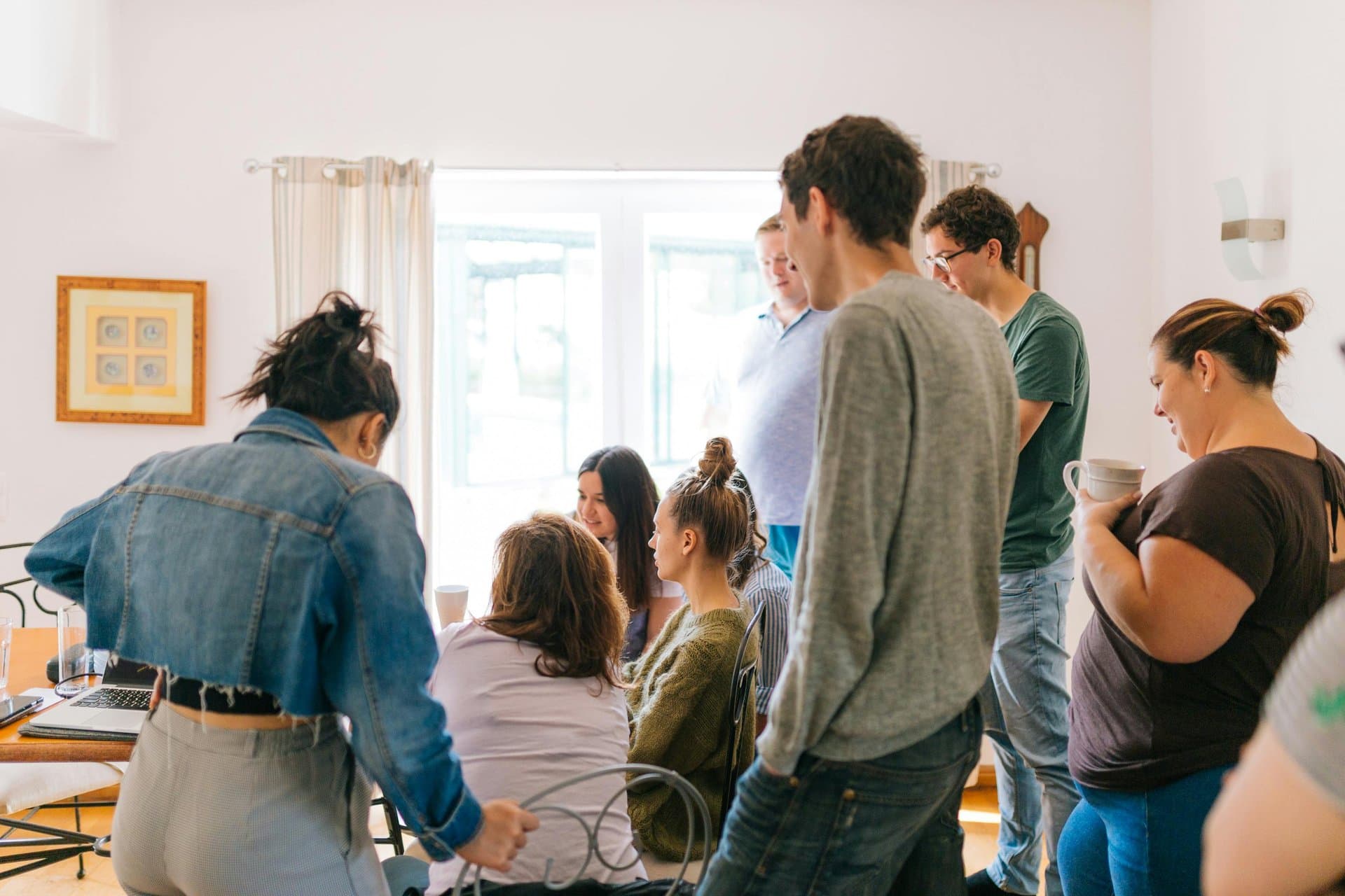 Diverse group of young adults gathered around a table in a sunlit room, talking and collaborating in a casual home setting.