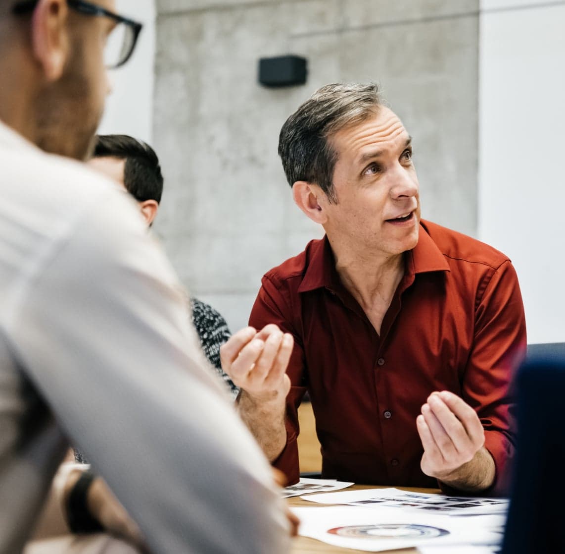 Three colleagues at a table; a man in a red shirt gestures while discussing printed charts