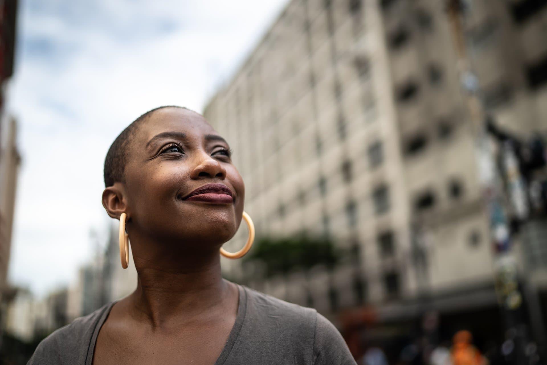 Smiling woman with short hair and gold hoop earrings looking upward hopefully on a city street with buildings in the background.