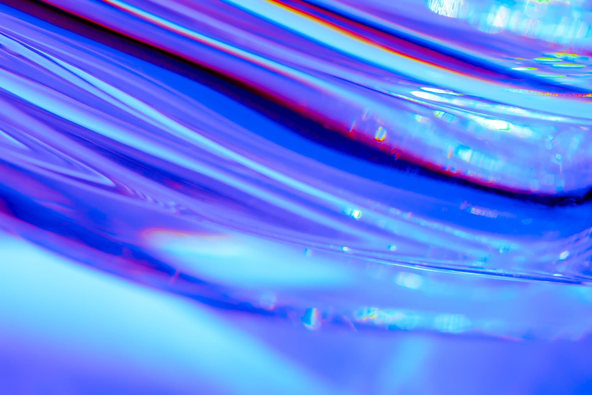 Abstract macro photograph of refracted light through blue glass creating flowing translucent waves and prismatic color highlights.
