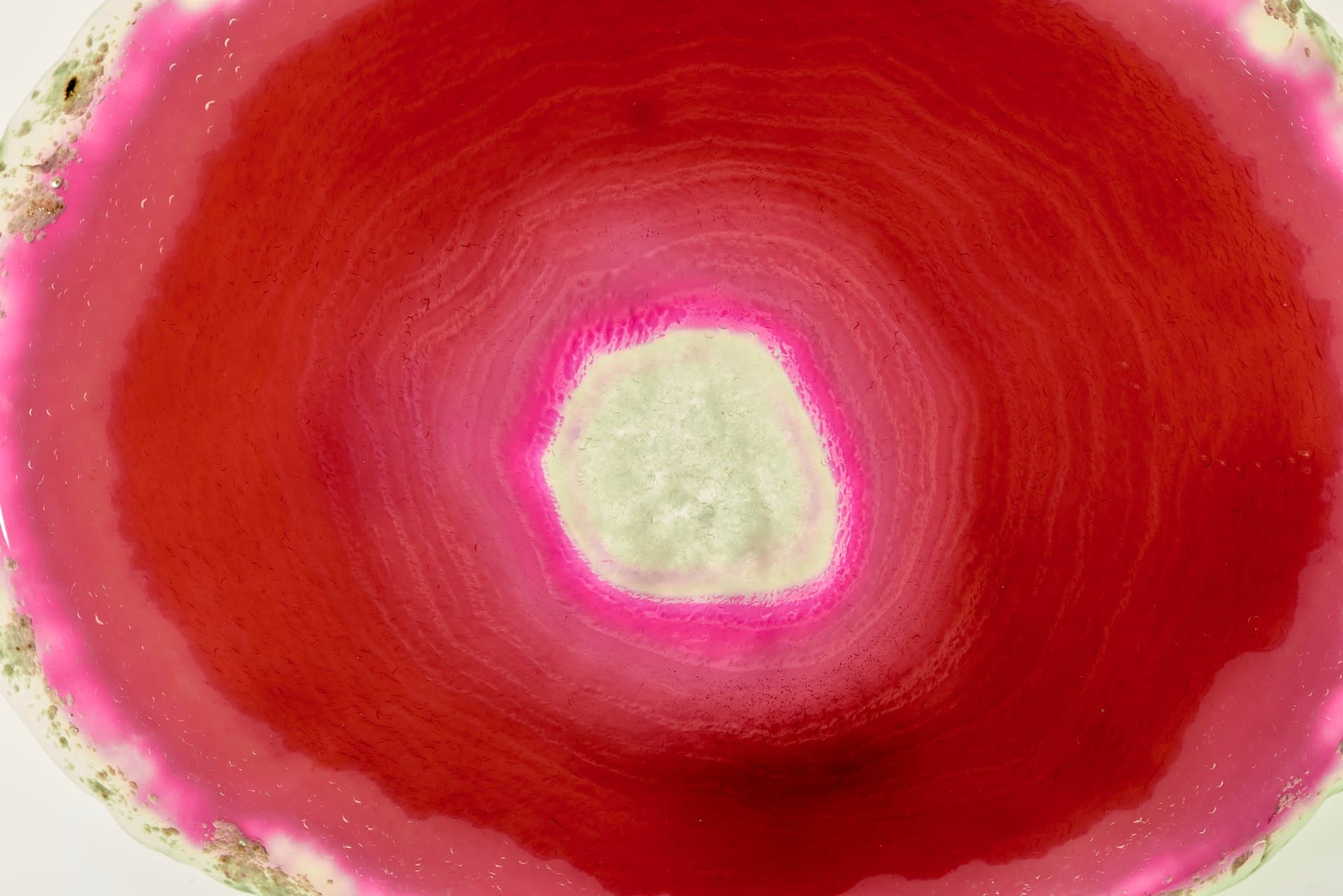 Vivid pink and red abstract image evoking a magnified biological cell or tissue sample with a pale green nucleus-like center.
