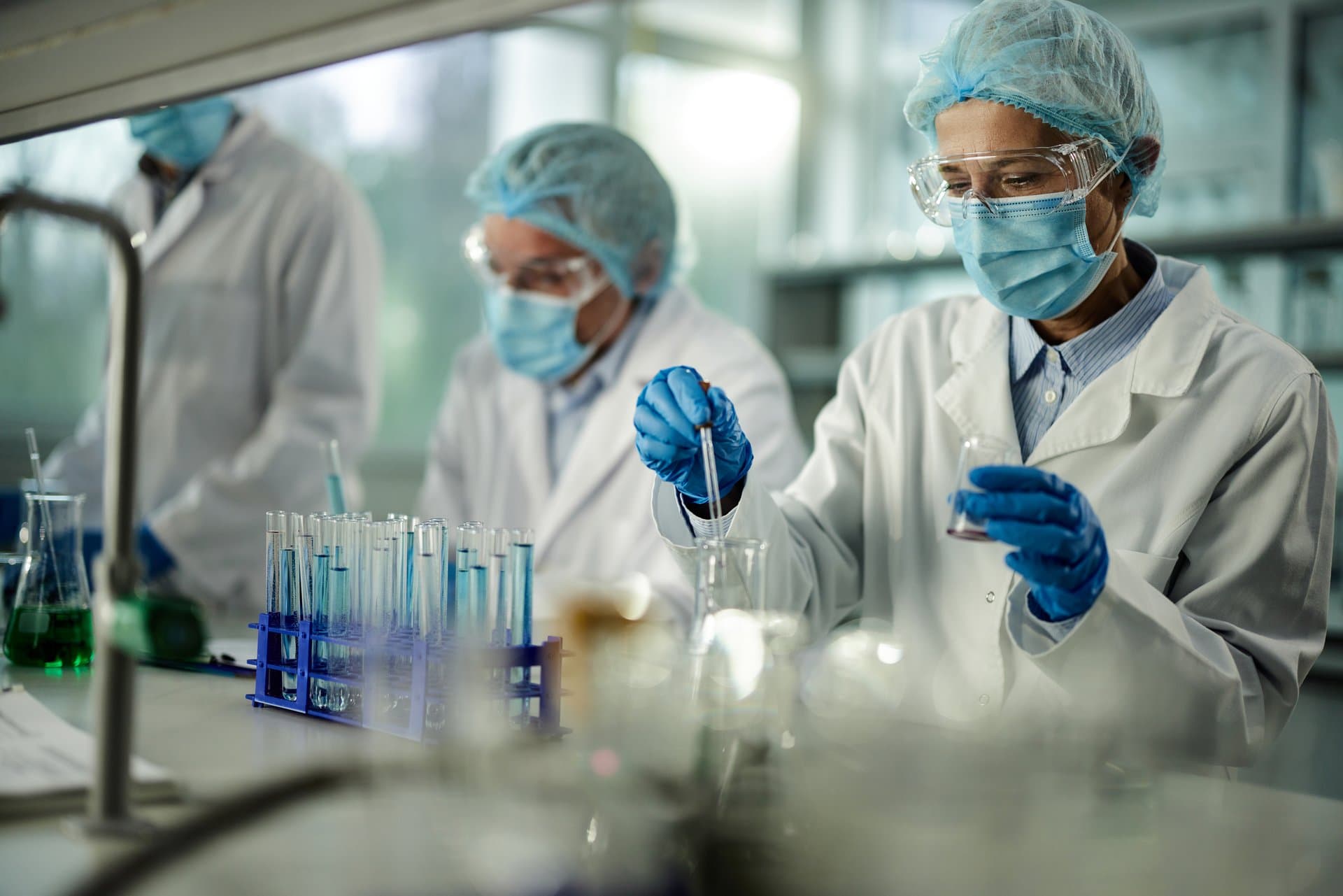 Female scientist in protective lab gear pipetting samples while colleagues conduct research with test tubes in a healthcare laboratory.